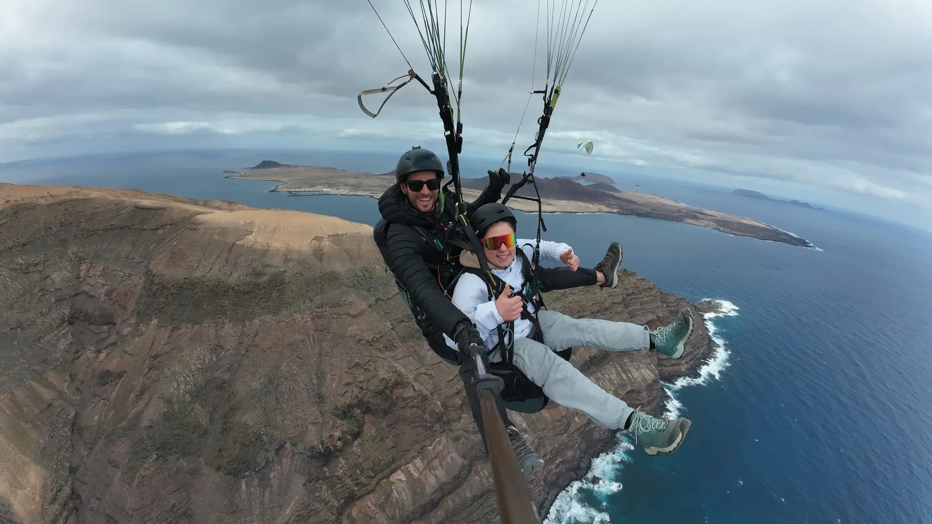 Smiling passenger giving a thumbs up on a tandem flight above the Famara cliffs, with La Graciosa behind